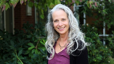 Woman with long grey hair and purple shirt smiling and standing outside