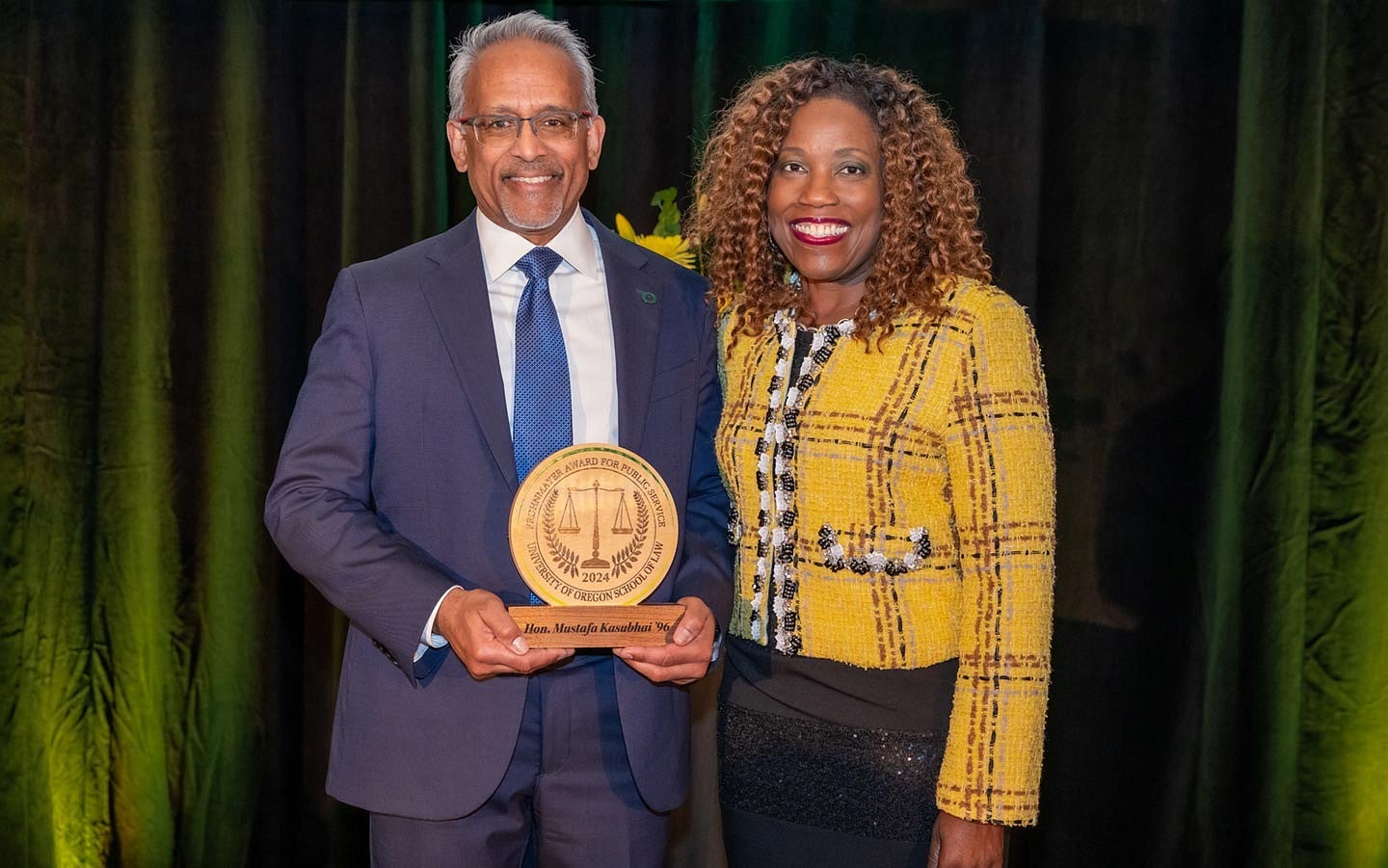 Judge Mustafa T. Kasubhai holding the Frohnmayer Award, standing next to Dean Marcilynn Burke in front of a green curtain.