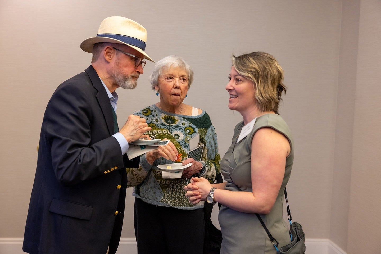 A man and two women speaking to each other while eating hors d'oeuvres with a white wall behind them.