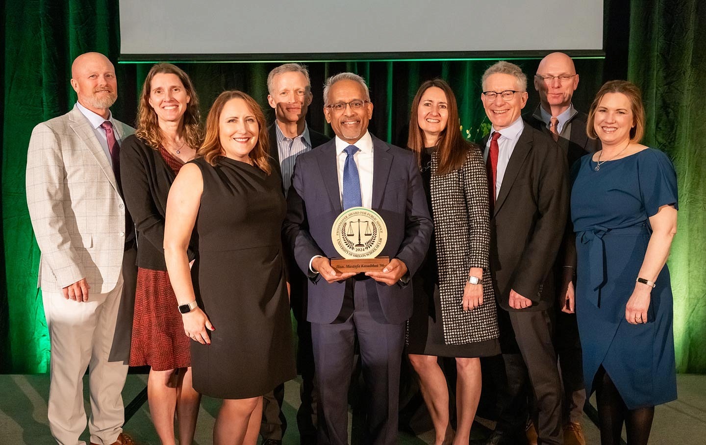 The University of Oregon School of Law class of 1996 posing with Judge Mustafa T. Kasubhai in the center holding the Frohnmayer award in front of a green curtain.