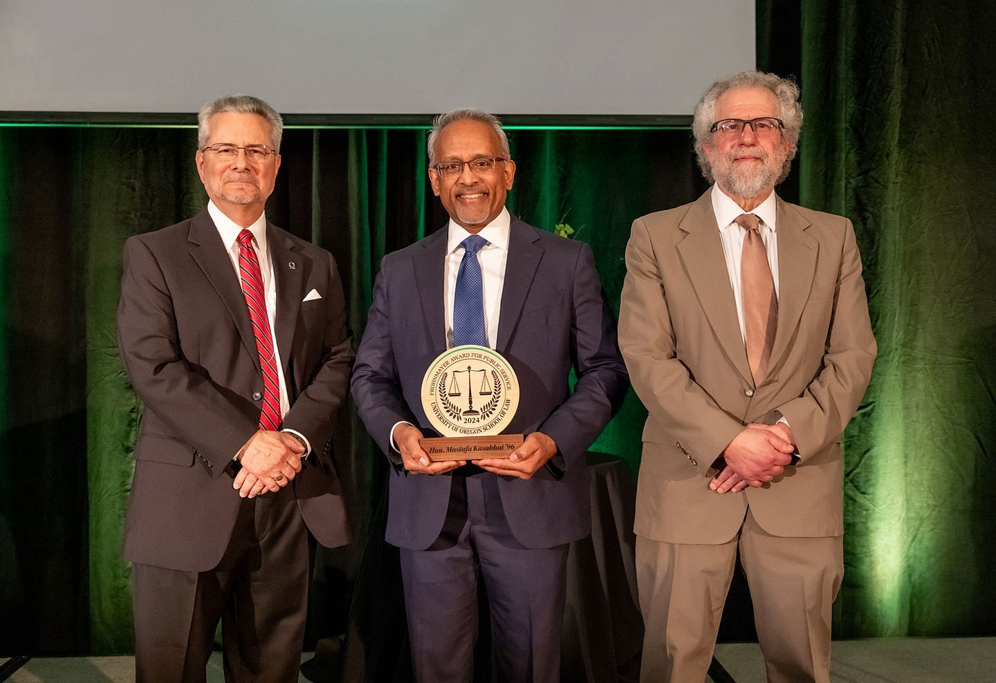 Judge John Acosta, Judge Mustafa T. Kasubhai holding the Frohnmayer Award, and attorney Marc Friedman standing in front of a green curtain.