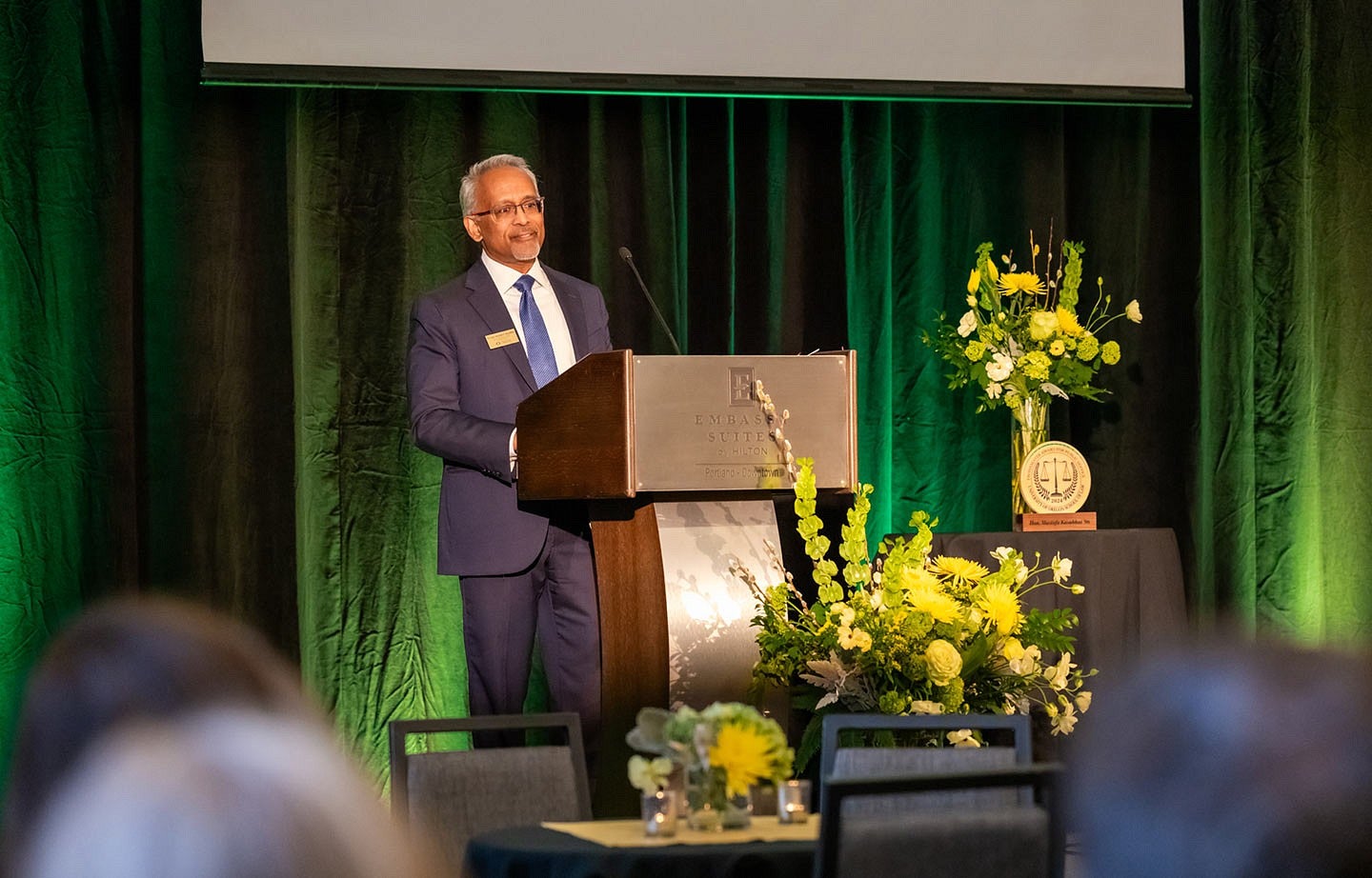 Judge Mustafa T. Kasubhai speaking at a podium on stage before an audience with a green curtain behind him for the Frohnmayer Awards ceremony.