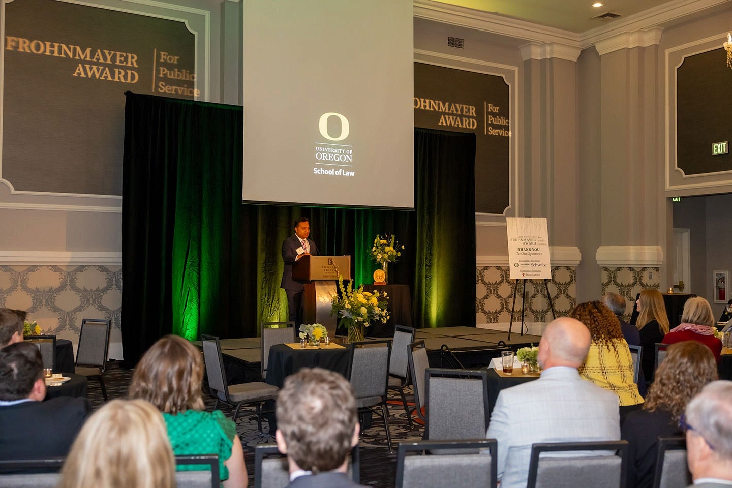 Abid Hussain speaking at a podium on stage before an audience with a green curtain and a projection screen behind him that reads "University of Oregon School of Law" below the yellow O logo at the Frohnmayer Awards.