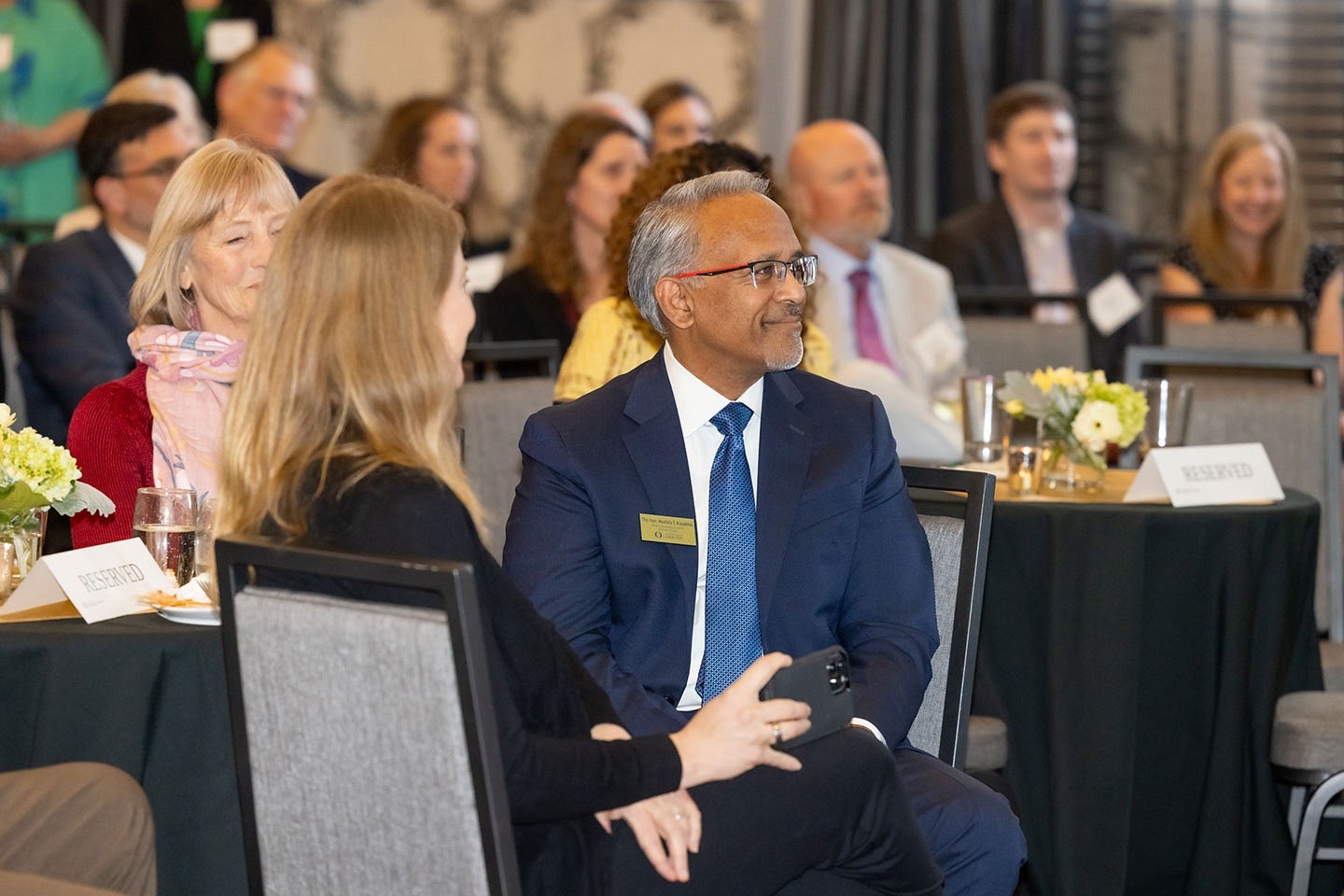 Judge Mustafa T. Kasubhai smiling and watching the Frohnmayer Awards ceremony from the crowd, seated at a table in a crowded ballroom.