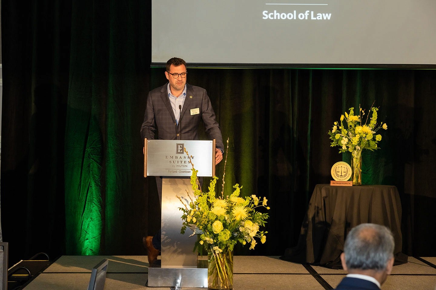 Matthew Schroettnig speaking at a podium on stage before an audience for the Frohnmayer Awards with a green curtain and projection screen that reads "School of Law" behind him.