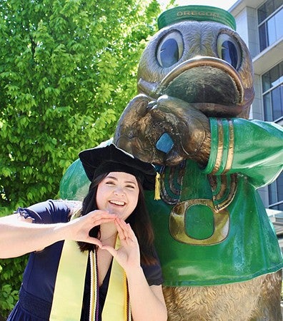 A student wearing graduation regalia posing with her hands in an "O" shape next to a statue of the University of Oregon duck mascot making the same pose outside of the Ford Alumni Center.
