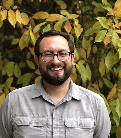 A photo of a smiling student with glasses and a beard standing in front of a bush.