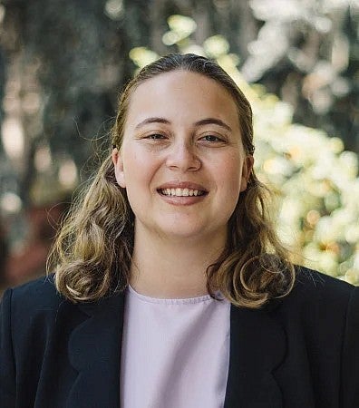 A photo of a student smiling in a pink shirt with a black blazer standing outside.