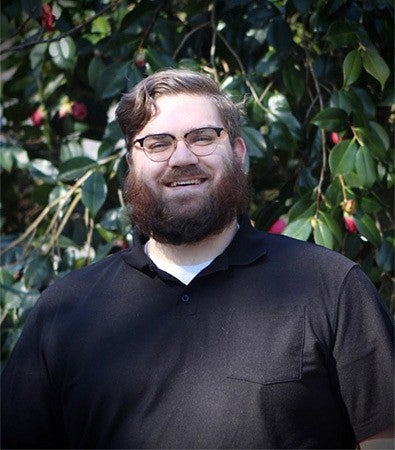 A student with glasses and a beard smiling and standing outside in front of a bush.