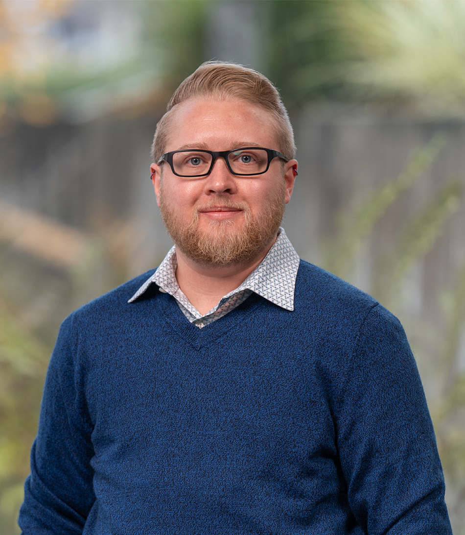 A man with blond hair and a beard wearing black square glasses and a navy blue sweater.