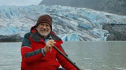 Professor Emeritus John Bonine wears a red jacket and hat and smiles while paddling a kayak