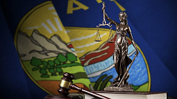 A gavel, textbook, and lady justice figurine are in focus before a blurred background of the Montana state flag