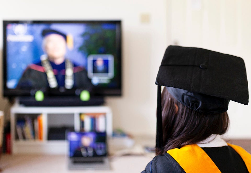 graduate in robe and cap watching commencement on tv in their livingroom.