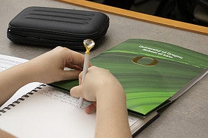 Student taking notes with a pen topped by a diamond-gem solitaire next to a University of Oregon School of Law folder