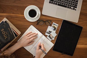 person holding ballpoint pen writing on a notebook with a laptop and coffee