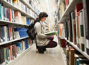 Student kneeling in a library aisle looking at an open book
