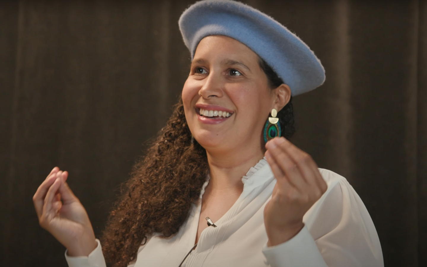 a woman with long brown curly hair and a grey beret smiling and gesturing with her hands