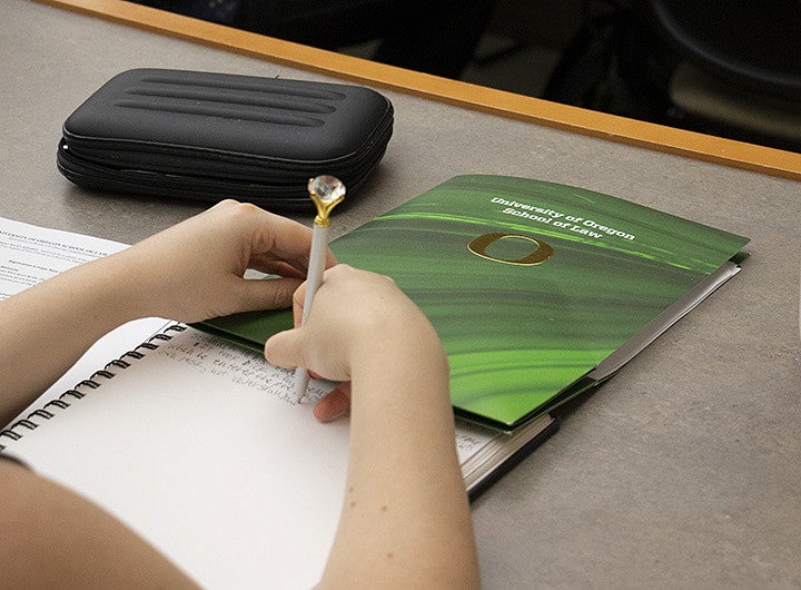 Student taking notes with a pen topped by a diamond-gem solitaire next to a University of Oregon School of Law folder