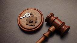 a generic silver key attached to wooden house cut-out keyfob sitting on a block with a gavel laying next to it